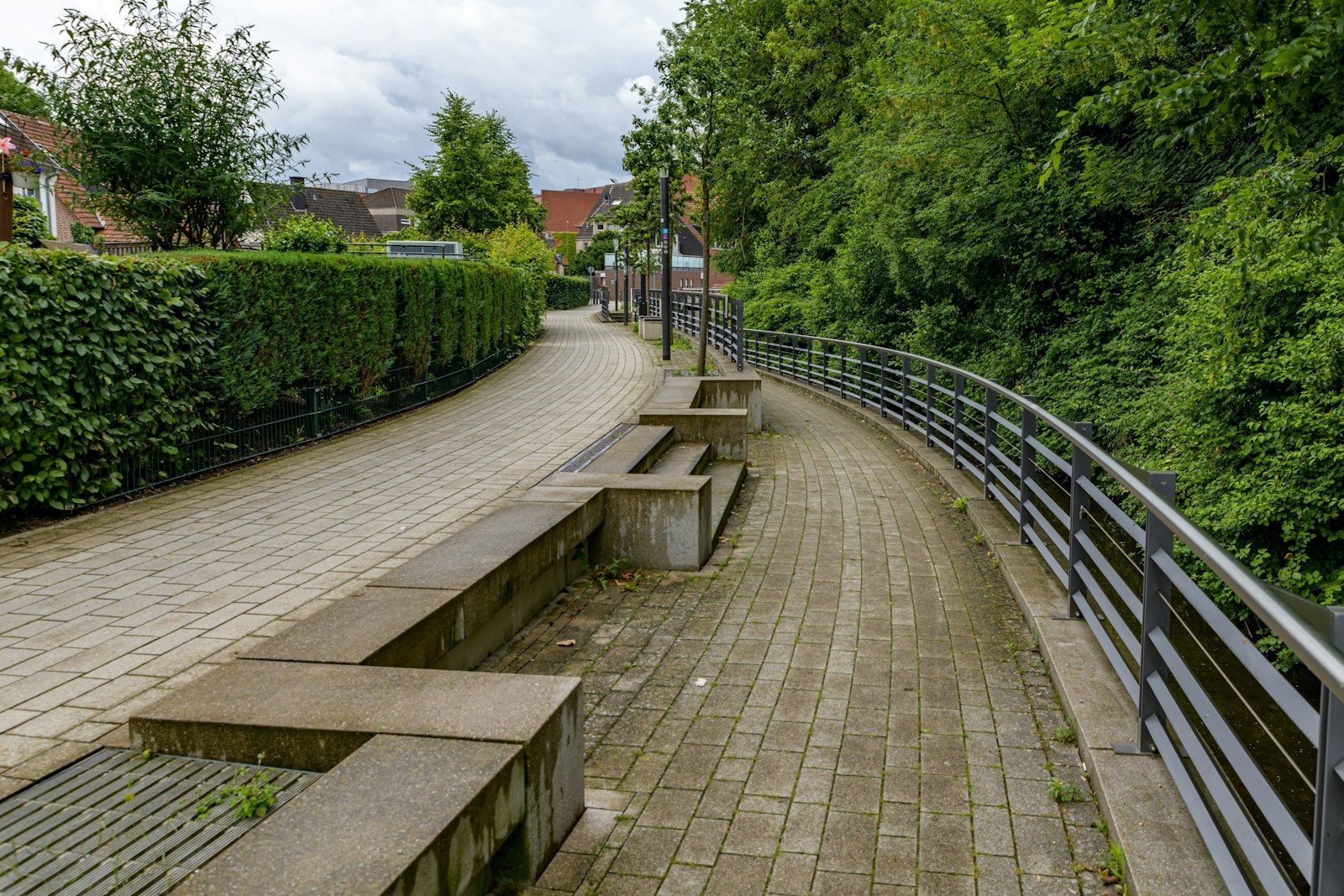 Outdoor walkway with stone benches, railing, and lush greenery.